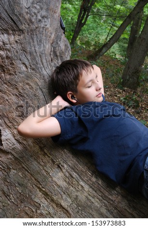 Teenage boy is resting with closed eyes. He is lying on the trunk of old oak. His hands are put behind the head. He is wearing T-shirt and jeans.