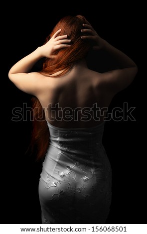 A young woman is standing back in the dark. She looses her long red hair. She is wearing only transparent nightdress.