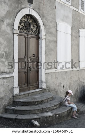 A modern preteen girl is waiting under the closed door. She is sitting on the stone stairs of the old house.