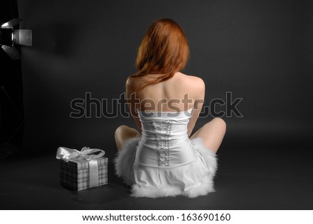 Girl sitting back near box gift in lotus position, dark background, white fancy dress decorated with down