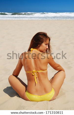 Young woman is sitting back on the sand sunlit beach. She is wearing yellow bikini. She is acquiring a tan in the sun. In the background there are sea waves under the clear sky.