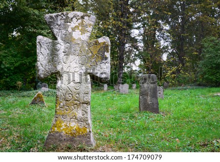 Ancient tombstones are in the green grass of old graveyard. In the foreground the large stone cross is covered with moss and antique epitaph.