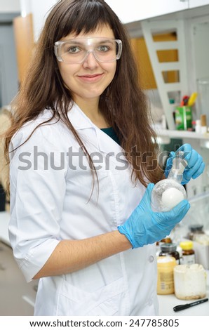 A young researcher holds a glass-wear with a substance in the chemical laboratory. She is wearing an overall, protective gloves and glasses.