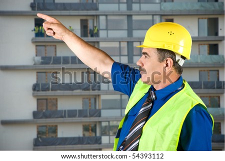 Single mature architect man pointing on site and looking up,construction with workers in background