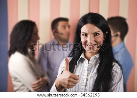 Happy customer service rep smiling and giving thumbs up in front of image with her team standing in background and having an conversation