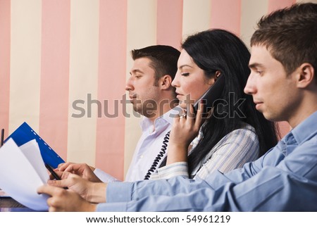 Group of business people sitting at desk in office and working,one businesswoman speaking at phone and her colleagues men reading papers,vertical blinds background