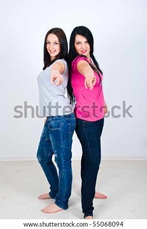 Two happy women in casual clothes standing back to back and pointing to you