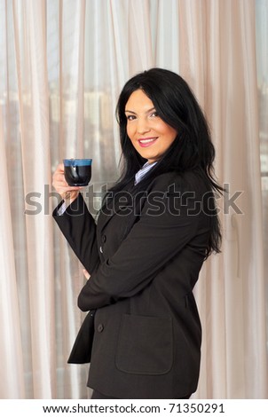 Smiling business woman standing near window in office and drinking a cup of coffee