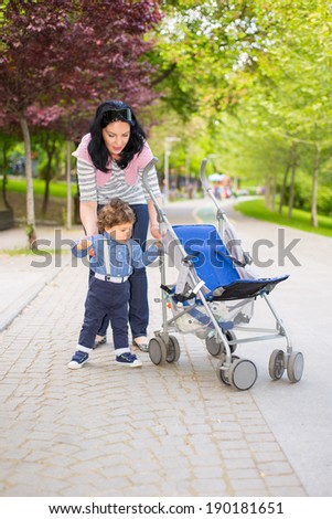 Mother helping toddler boy to pushing  his pram in park