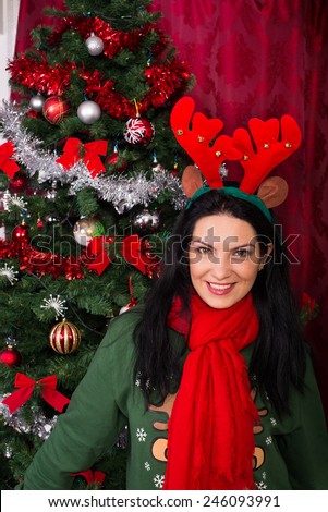 Smiling woman in red and green clothes wearing Reindeer ears and standing near Xmas tree