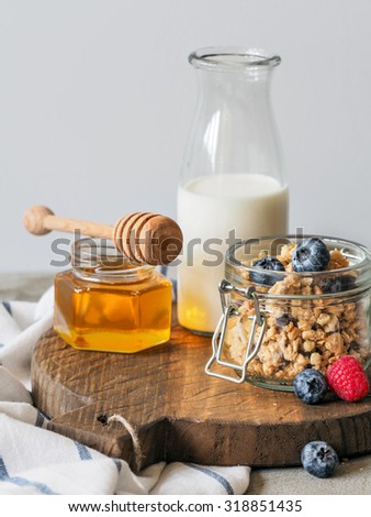 Homemade granola with blueberries and raspberries in open glass jar, bottle of milk and honey on rustic wooden cutting board