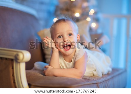 A lovely 3 year old girl in a beautiful white dress lying on a sofa in front of the decorated Christmas tree and laughing