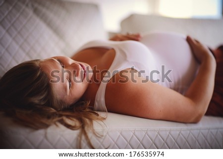 A portrait of a white beautiful young pregnant woman lying on a white sofa and touching her baby bump with a happy smile of a future mother