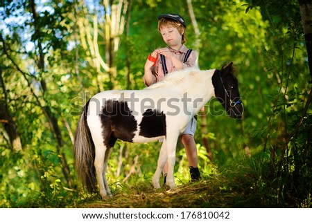 A cute white boy in a cap caressing his little pony in a green forest on a sunny summer day