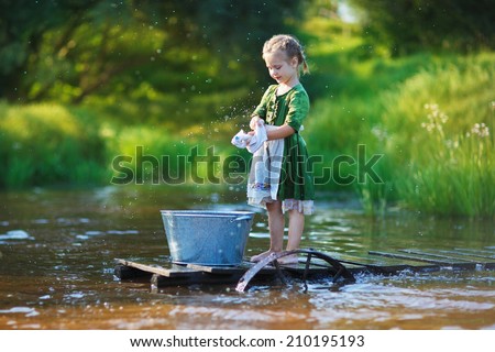 A cute little girl doing the laundry in a small aluminium basin standing on a pontoon at the pond in a sunny summer day. Kids are playing.