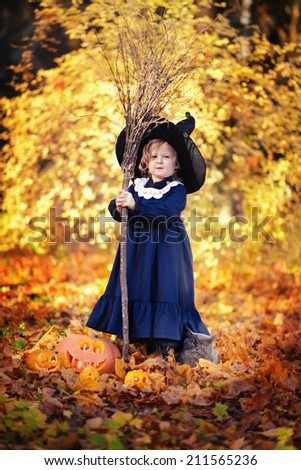 Little cute girl in black witch costume and a magic hat standing among orange leaves with a broom in a sunny autumn day. Halloween. National holidays and traditions. Fairy tale. Funny kids.