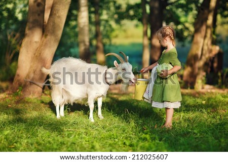 A little pretty blond girl in a green dress and white apron holding a milk-can and playing with her pet goat in the field in a sunny summer day. Kids as helpers in the country.