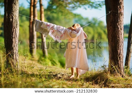 A little cute girl hanging washing on a line in a pine forest on the bank of a lake in a sunny summer day. Kids are playing.