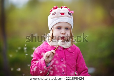 A pretty happy little girl in a pink jacket and white hat blowing a dandelion in cool weather in a forest. Kids and nature.