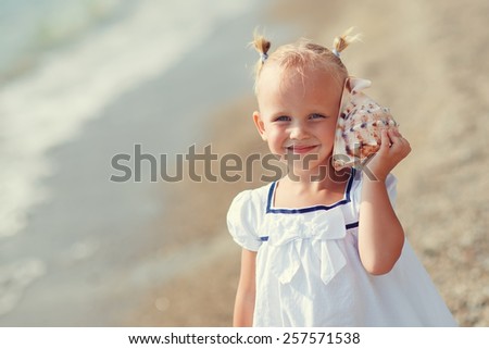 A portrait of a cute little smiling girl in white clothes playing with a shell on the beach on a warm sunny summer day. Holidays at sea. Funny kids