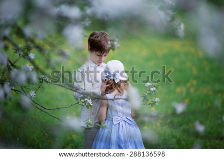 A cute little girl in an elegant white hat and light blue dress and a cute young boy embracing in the blooming garden on a sunny spring day. Kids in the country.