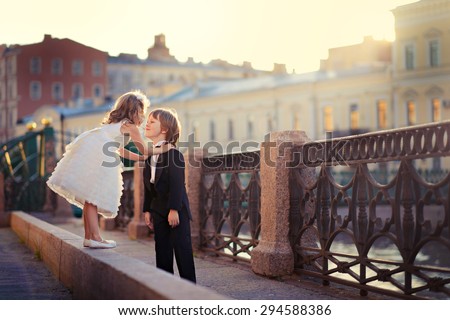 An elegant handsome little boy in a tail-coat and his pretty little girlfriend walking along a beautiful old bridge at sunset on a sunny warm summer evening. Kids in the city. Saint-Petersburg. Russia