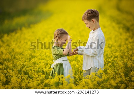 Little pretty girl in green Bavarian dress with white apron and young boy picking flowers in field of flowering yellow mustard on sunny summer day.Kids and nature.Children in country.Beautiful flora