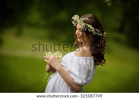 Happy motherhood: a young beautiful pregnant woman in a white dress and diadem of flowers walking in the park on a sunny summer day. Nature in the country.