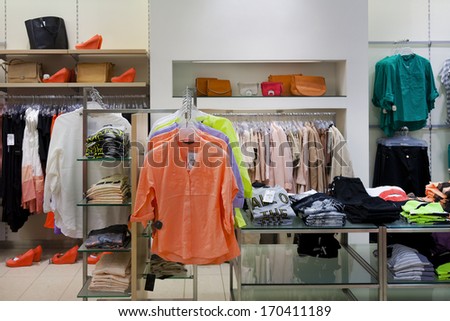 Blouses, shirts hanging on rack in retail shop interior. Shoes and handbags on shelf.