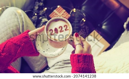 Number 2022 on frothy surface of cappuccino served in white cup holding by woman in red knitted sweater relaxed sitting on the couch. New year new you, Holidays food art theme Happy New Year 2022.