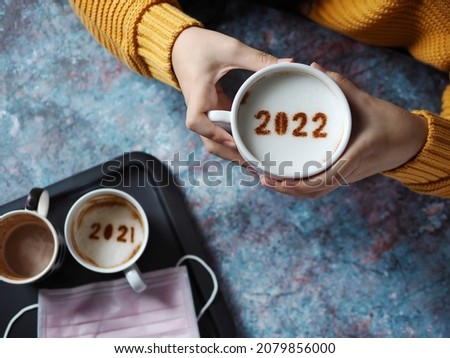 Hello 2022 Goodbye 2021 and COVID-19, female hands holding coffee cup with number 2022 on frothy surface, another one with 2021 at bottom placed on black aluminium tray with disposable medical mask.
