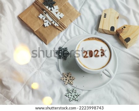 White coffee cup with number 2023 over frothy surface flat lay on the bed with white blanket, wooden house model, paper gift bag, snowflake sign. Home 2023, Happy new year theme (top view, copy space) Foto d'archivio © 