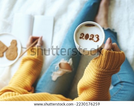 New Year's goal setting, number 2023 on frothy surface of cappuccino in white coffee cup holding by woman in yellow knitted sweater with jeans sitting on bed while writing down her resolutions. Foto d'archivio © 