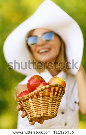 Smiling beautiful young woman in hat wiht wicker basket holds red apples, against green of summer park.