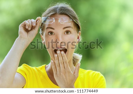 Dark-haired smiling beautiful young woman in yellow blouse and glasses is surprised, against green of summer park.