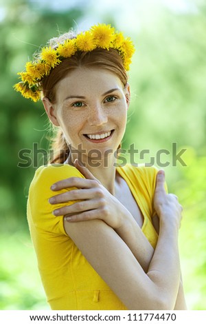 Portrait of red-haired smiling beautiful young woman in yellow blouse with wreath of dandelions, against green of summer park.