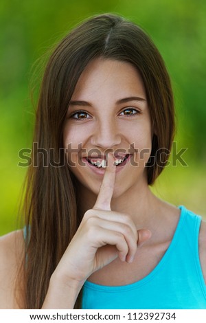 Close up of smiling beautiful teenage girl puts finger to his lips in token silence, against green of summer park.