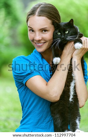 Dark-haired smiling beautiful young woman in blue blouse with black cat, against green of summer park.
