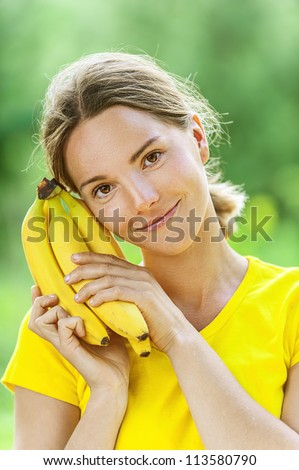 Portrait of dark-haired smiling beautiful young woman in yellow blouse with banana, against green of summer park.