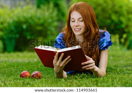 Red-haired smiling beautiful young woman in blue blouse lying on grass with book, against green of summer park.