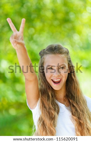 Beautiful teenage girl in white blouse up two fingers in victory sign, against green of summer park.