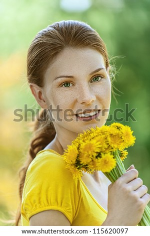 Portrait of red-haired smiling beautiful young woman in yellow blouse with bouquet of dandelions, against green of summer park.