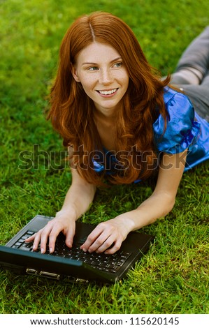 Red-haired smiling beautiful young woman in blue blouse lying on grass with laptop, against green of summer park.