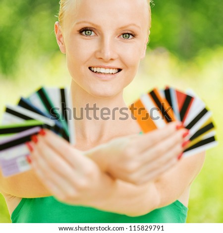 Portrait of pretty smiling fair-haired young woman holding cards at summer green park.