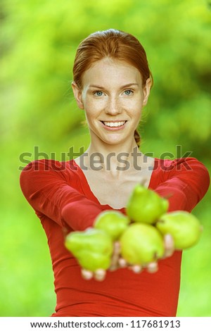 Portrait of red-haired smiling beautiful young woman in red blouse with pears, against green of summer park.