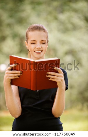 Beautiful smiling young woman in dark blouse reads red book, against green of summer park.