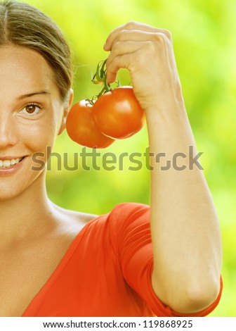 Portrait half of dark-haired smiling beautiful young woman in red blouse with tomato, against green of summer park.
