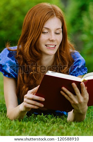 Red-haired smiling beautiful young woman in blue blouse lying on grass with book, against green of summer park.
