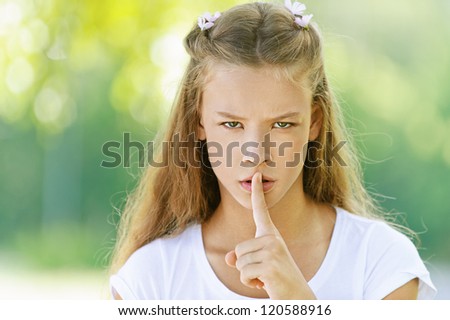 Beautiful teenage girl in white blouse raised index finger to her lips, against green of summer park.