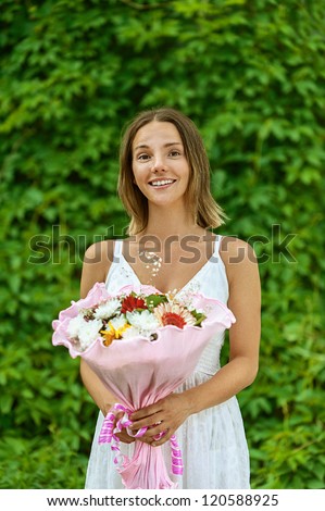 Dark-haired smiling beautiful young woman in white blouse with bouquet of flowers, against green of summer park.